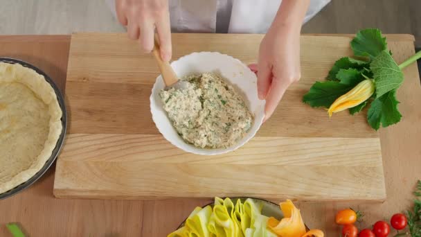 A girl chef prepares a curd filling with herbs, puts it on the pie dough in a baking dish, on a wooden table, with zucchini vegetables and tomatoes in the background. Close-up, top view. - Footage, Video