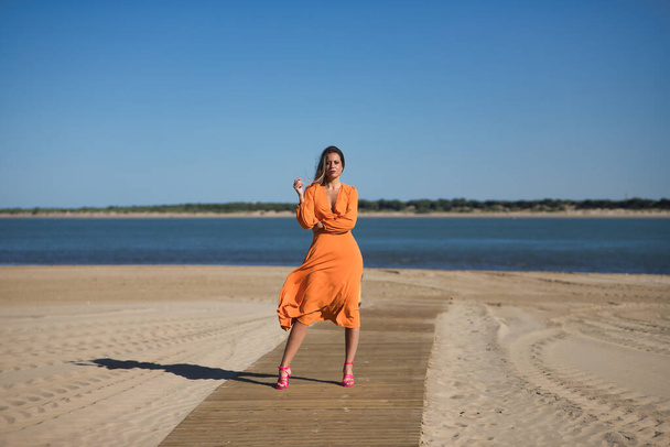 Bella giovane donna in un abito arancione, in posa su una passerella di legno sulla spiaggia, con il vento arruffando i capelli e il vestito. Concetto bellezza, moda, tendenza, aria, spiaggia. - Foto, immagini
