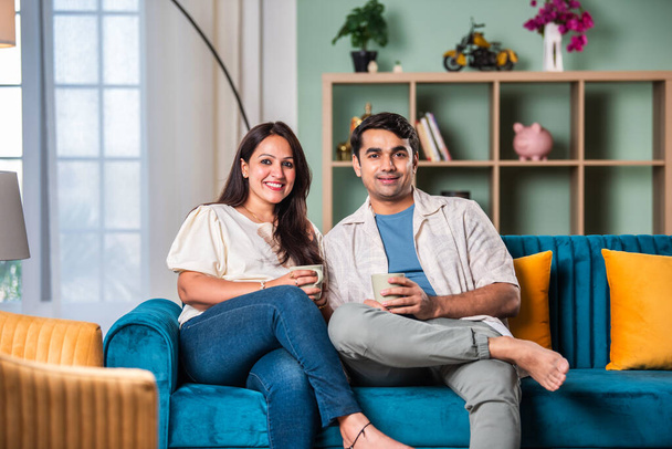 Щаслива індійська молода пара має чай або каву разом, сидячи на дивані вдома - Фото, зображення