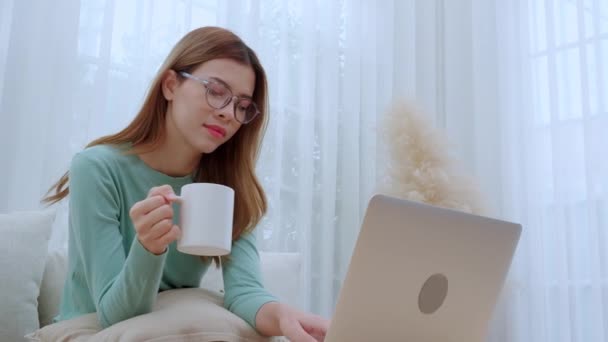 Jonge Aziatische vrouw met een bril zitbank werkend op laptop computer en het drinken van koffie in de woonkamer thuis, vrouw werk van thuis met telewerk, freelance werken, het bedrijfsleven en het digitale leven. - Video
