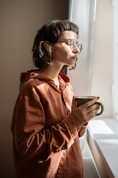 Страшная женщина-интроверт в очках смотрит в окно и пьет чай дома. Девушка, страдающая психическими заболеваниями, смотрит на людей, машины, думает о жизненных проблемах. Одиночество.  - Фото, изображение