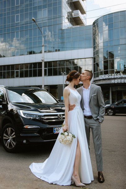 novia y novio con un coche negro cerca de un rascacielos de cristal - Foto, imagen