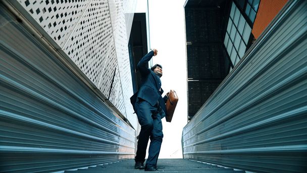 Low angle view of young smiling business man dance between building at center point. Skilled executive manager wearing suit and suitcase and moving to music. Happy investor moving to music. Exultant. - Photo, Image