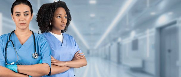 Twee serieuze Europese, Afrikaanse Amerikaanse verpleegsters in blauwe operatiekleding met gekruiste armen en stethoscopen staan vastberaden in een ziekenhuisgang, die kracht en professionaliteit weerspiegelen. - Foto, afbeelding