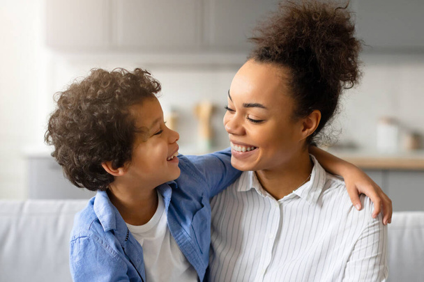 Affectueuse jeune mère noire avec un large sourire embrasse son fils aux cheveux bouclés, profitant tous deux d'un moment chaleureux et aimant dans un salon confortable et ensoleillé - Photo, image
