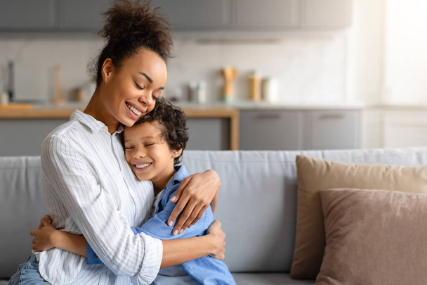 Vreugdevolle zwarte tiener jongen in warme omhelzing met zijn liefhebbende moeder, stralend geluk en familiale liefde in gezellige huiselijke omgeving, samen zitten op de bank - Foto, afbeelding