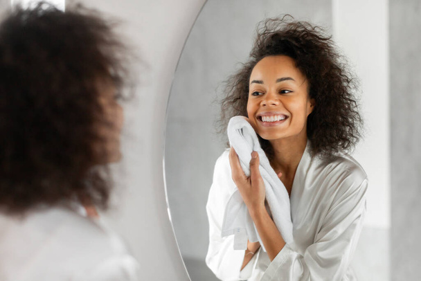 Cuidado de la piel facial e higiene. Pretty Black Lady secando la cara limpia con una toalla suave después de lavarse en el baño, sonriendo a su reflejo en el espejo, enfoque selectivo. Rutina de belleza matutina - Foto, Imagen