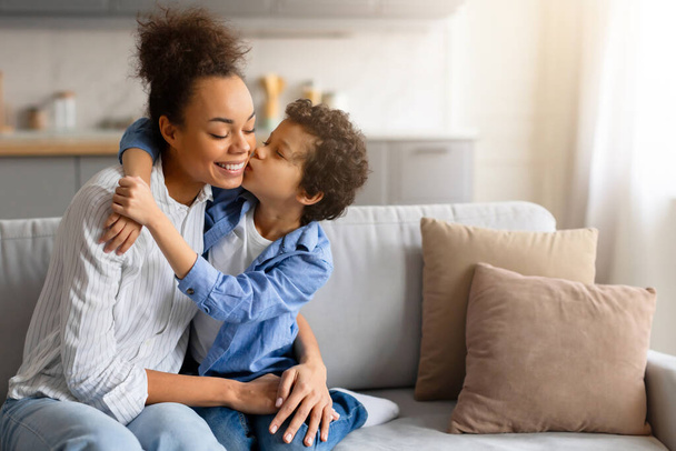 Preteen garçon noir en chemise bleue embrassant affectueusement sa joue de mères heureuses, mettant en valeur leur relation d'amour dans un cadre confortable à la maison, espace libre - Photo, image