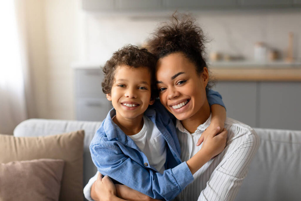 Η λαμπερή μαύρη μητέρα και ο νεαρός γιος της μοιράζονται εγκάρδια αγκαλιά, τα πρόσωπά τους λάμπουν από ευτυχία, σε ένα άνετο σπιτικό περιβάλλον που ακτινοβολεί οικογενειακή ζεστασιά. - Φωτογραφία, εικόνα