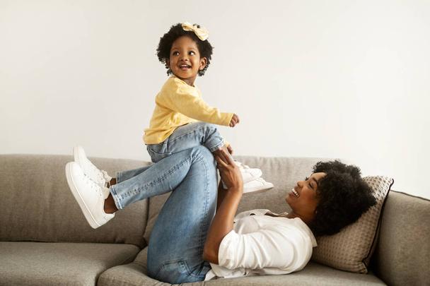 Side näkymä onnellinen afrikkalainen amerikkalainen nuori äiti ja pieni tyttö leikkii yhdessä sohvalla kotona sisustus, kopioi tilaa. Äiti nauttii vauvalapsensa kanssa. Äitiyden käsite - Valokuva, kuva