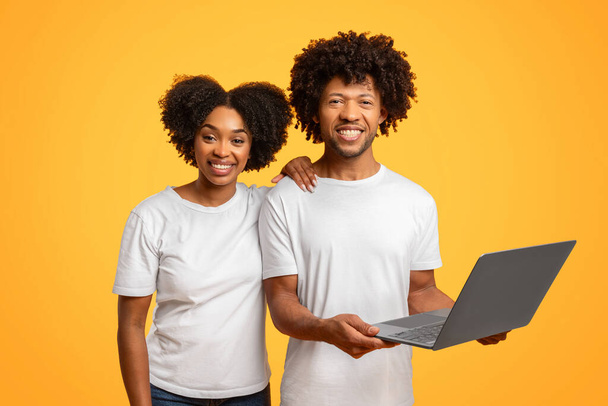 Sonriendo fresco millennial africano americano pareja con camisetas blancas usando ordenador portátil en colorido estudio fondo, compras o la banca juntos, asistir a curso en línea juntos - Foto, Imagen