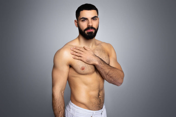 Sexy topless middle eastern young man shirtless looking at camera, taking care of his body, touching his chest, skincare after shower, isolated in grey background, copy space - Фото, изображение
