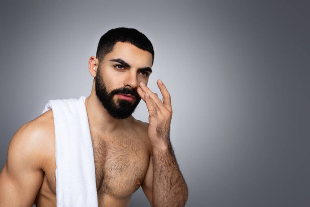 Όμορφος γενειοφόρος χιλιετής γυμνός Άραβας με πετσέτα μπάνιου στον ώμο απλώνοντας κρέμα στο πρόσωπο, κοιτάζοντας το χώρο αντιγραφής που απομονώνεται σε γκρι φόντο στούντιο. ρουτίνα φροντίδας του δέρματος για τους άνδρες - Φωτογραφία, εικόνα