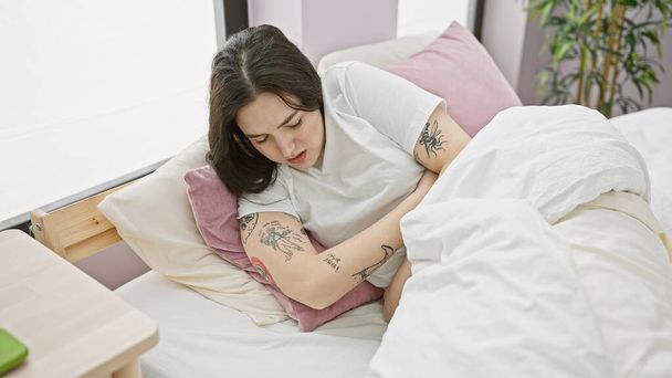 Молодая татуированная белая женщина страдает от боли, сжимая желудок, сидя дома в постели.. - Фото, изображение