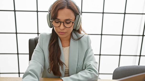 Captivating portrait of a young, beautiful hispanic woman, immersed in her work at the office, showcasing an elegant business look, listening to soothing music through her headphones - Foto, immagini