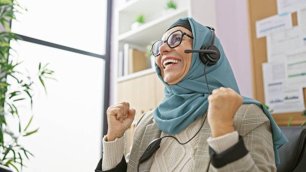 A jubilant, middle-aged woman in a hijab with a headset celebrates in an office environment, exemplifying diversity and success at work. - Photo, Image