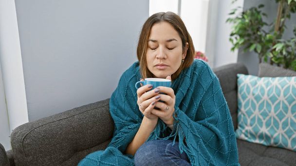 A relaxed woman enjoys a warm drink on a sofa in a cozy living room, wrapped in a teal blanket. - Photo, Image