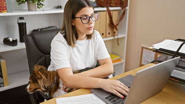 Eine fokussierte junge Frau arbeitet an einem Laptop in einem modernen Büro, ihren treuen Hund an ihrer Seite - Foto, Bild