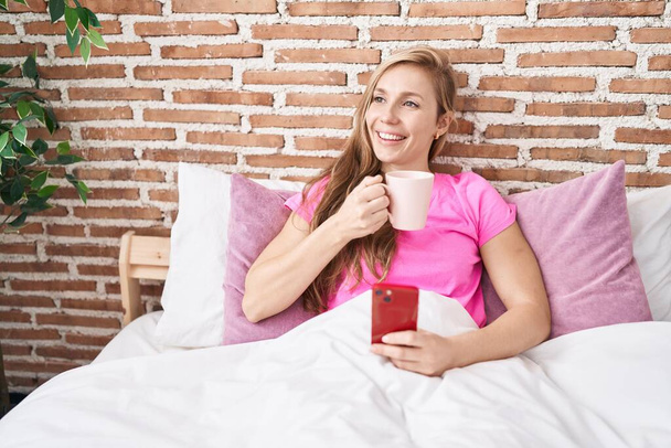 Young blonde woman using smartphone drinking coffee at bedroom - Foto, imagen
