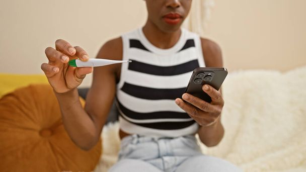 Donna afro-americana preoccupata a casa, che prende la temperatura con un termometro, incollata allo schermo del suo smartphone - teleconsultante, messaggistica in mezzo alla febbre - Foto, immagini
