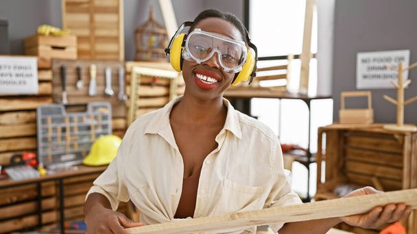 Hermosa mujer afroamericana carpintera, adornada con gafas de seguridad y auriculares, manejando magistralmente tablón de madera en el bullicioso taller de carpintería - Foto, Imagen