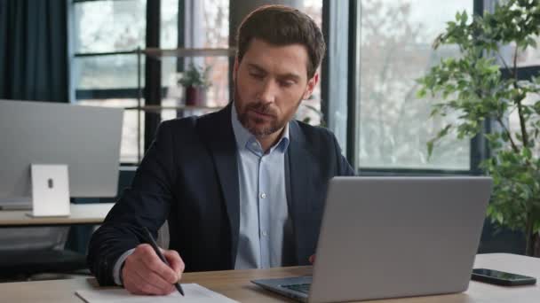 Kaukázusi férfi üzletember szakmai iroda alkalmazott HR manager vállalkozó Vezérigazgató írni adatokat beszél videohívás segítségével laptop a munkahelyen online üzleti konferencia távoli tárgyalások webinar - Felvétel, videó