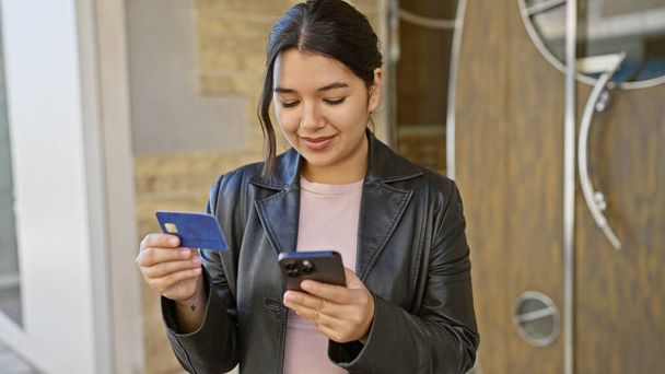 Egy fiatal spanyol nő ellenőrzi a hitelkártyáját és az okostelefonját egy városi utcában, megtestesítve a városi eleganciát és kapcsolódást.. - Fotó, kép