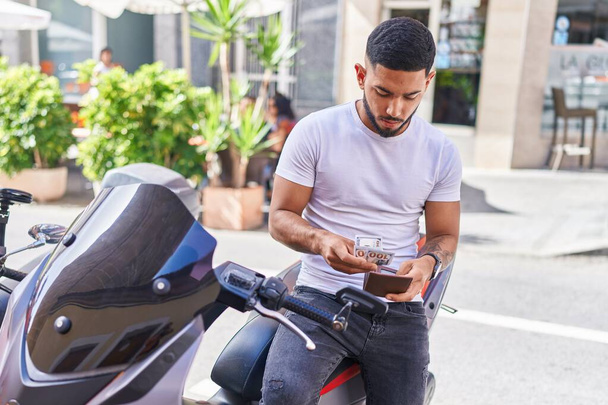 Молодий латинський чоловік рахує долари, сидячи на мотоциклі на вулиці
 - Фото, зображення
