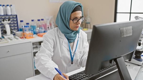 Una mujer madura en un hiyab trabajando en una computadora en un laboratorio - Foto, Imagen