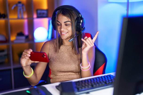 Νεαρή ισπανόφωνη γυναίκα που παίζει βιντεοπαιχνίδια με το smartphone χαμογελώντας χαρούμενη δείχνοντας με το χέρι και το δάχτυλο στο πλάι  - Φωτογραφία, εικόνα
