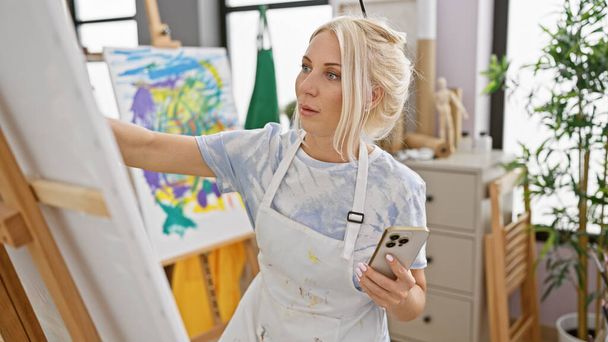 In uno studio d'arte rilassato, una giovane, attraente artista bionda, immersa nella sua creatività, guardando il suo smartphone, disegna abilmente, dando vita alla sua tela.. - Foto, immagini