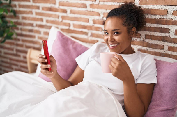 Αφροαμερικανή γυναίκα που χρησιμοποιεί smartphone πίνοντας καφέ στο υπνοδωμάτιο - Φωτογραφία, εικόνα