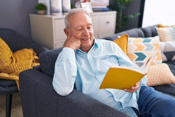 Homem de cabelos grisalhos de meia idade lendo livro sentado no sofá em casa - Foto, Imagem