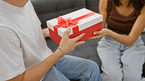 Egy férfi ajándékoz egy nőnek egy piros dobozt masnival egy kényelmes nappaliban, kifejezve szeretetét és meglepetését.. - Fotó, kép