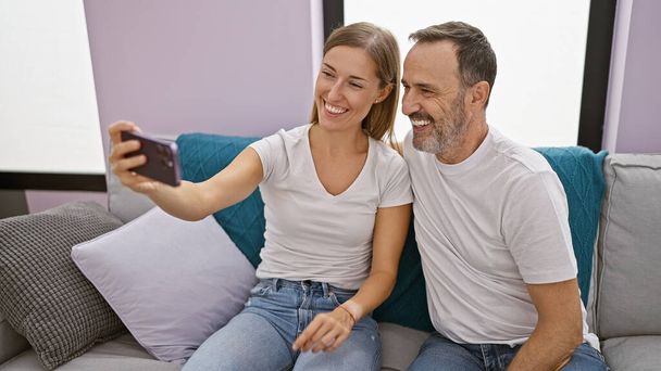 Glückliche Vater und Tochter, die innige Erinnerungen wecken, selbstbewusst lächeln, während sie auf dem Sofa sitzen und zu Hause ein entspanntes Selfie mit dem Smartphone machen. - Foto, Bild