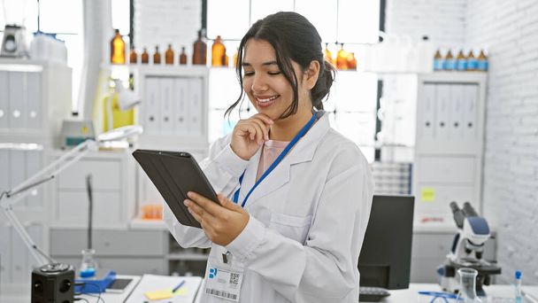 Hymyilevä nuori latinonainen laboratoriotakissa käyttää tablettia nykyaikaisessa laboratoriossa, joka edustaa lääketieteellistä ammattitaitoa ja teknologiaa.. - Valokuva, kuva