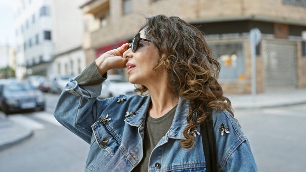 Зріла іспаномовна жінка з кучерявим волоссям коригує свої сонцезахисні окуляри, стоячи на відкритому повітрі в міському містечку. - Фото, зображення
