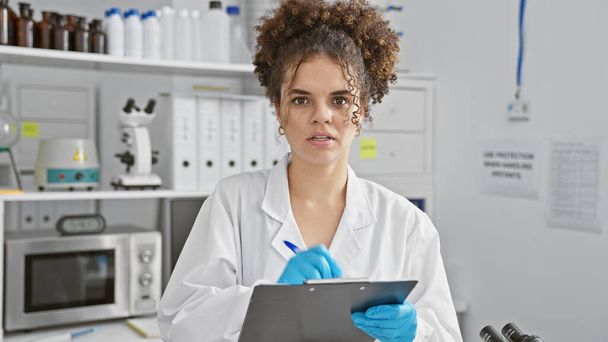 Una giovane donna ispanica dai capelli ricci, vestita con camice e guanti, prende appunti in laboratorio. - Foto, immagini