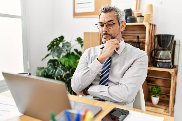 Середнього віку сіроволоса людина бізнесмен використовує ноутбук з сумнівом експресія в офісі
 - Фото, зображення