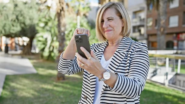 Usmívající se starší žena používá smartphone ve slunečném parku - Fotografie, Obrázek