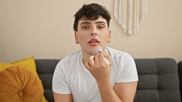 Komea nuori mies käyttää meikkiä harjalla modernissa olohuoneessa, jossa esitellään sukupuolen sujuvuutta ja itsehoitoa kotona. - Valokuva, kuva