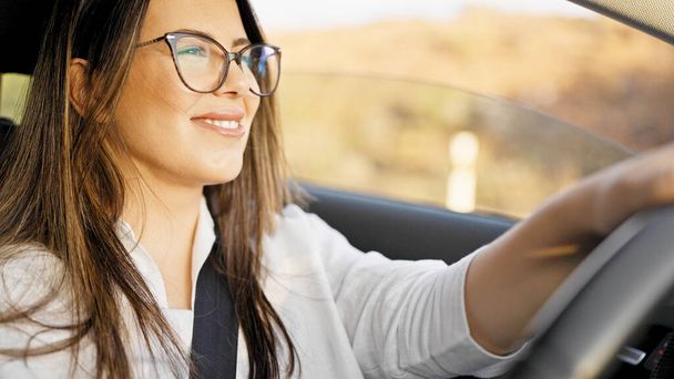 Mladý krásný hispánec žena řízení auto s úsměvem nosí brýle na silnici - Fotografie, Obrázek