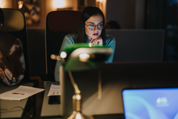 Uma jovem profissional dedicada está imersa em seu trabalho no escritório durante as horas da noite, mostrando determinação e produtividade.. - Foto, Imagem
