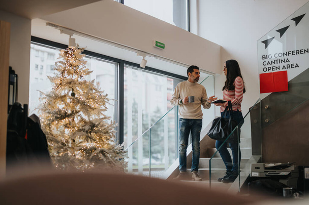 Dos colegas conversando mientras sostienen tazas de café junto a un árbol de Navidad decorado festivamente en un entorno de oficina moderno. - Foto, imagen