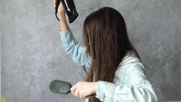Девушка сушит влажные волосы феном, используя расческу для волос на сером фоне, вид сбоку. Идея ухоженных волос. Высококачественные 4k кадры - Кадры, видео