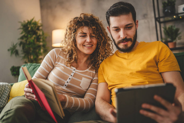 muž a žena kavkazský dospělý pár číst knihu a používat digitální tablet doma na rozkládací pohovce - Fotografie, Obrázek