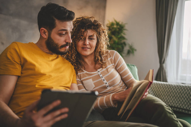 男と女性の慎重な大人のカップルは,ソファベッドで自宅で本を読み,デジタルタブレットを使用しています - 写真・画像