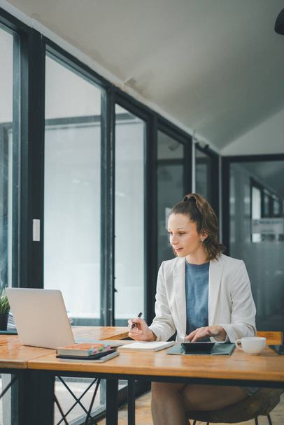Business Analytics-Konzept: Eine Geschäftsfrau arbeitet konzentriert an ihrem Laptop in einem hellen Büro mit Kaffeetasse und Smartphone auf dem Schreibtisch. - Foto, Bild