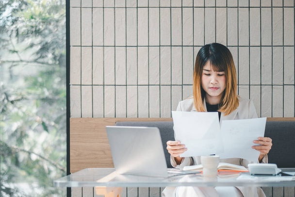 Conceito freelancer, Empresária focada em um blazer bege revisa papelada com seu laptop em uma mesa de café, incorporando multitarefa profissional - Foto, Imagem
