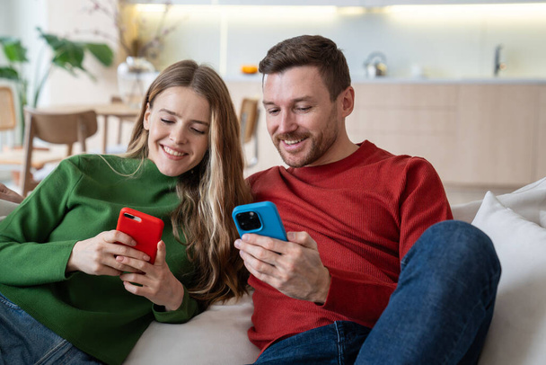 Fröhliches glückliches Paar, das mit Smartphones auf dem Sofa sitzt, lächelt, fesselndes Online-Spiel spielt, kurze lustige Videos im Internet anschaut, Fotos austauscht, Gruppenvideoanrufe mit Freunden führt - Foto, Bild